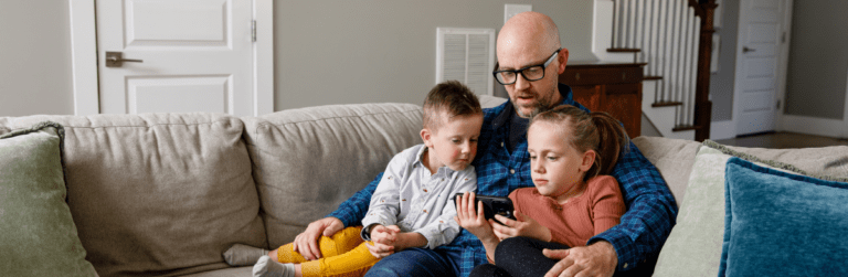 Father sitting on couch with son and daughter, all looking at cell phone screen the daughter is holding