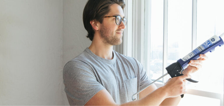 A homeowner uses a caulking gun to apply sealant along a residential window frame to reduce drafts and improve home air sealing.