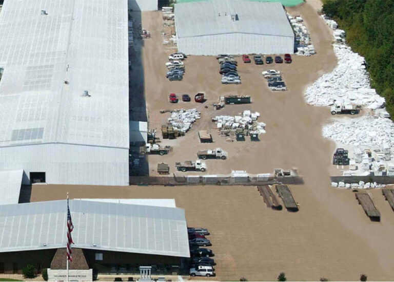 Aerial view of a large industrial manufacturing facility featuring white warehouse roofs, outdoor supply yards, and employee parking areas.