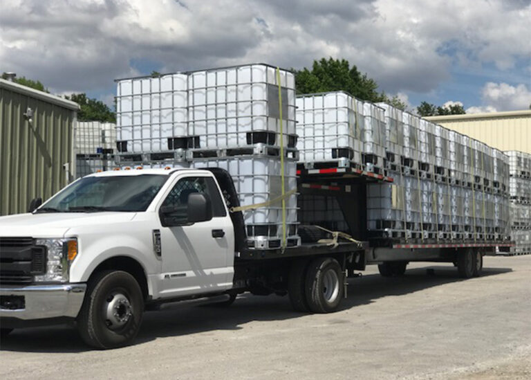 A white commercial work truck pulling a long flatbed trailer loaded with multiple stacked intermediate bulk container (IBC) totes for industrial liquid management and facility operations.