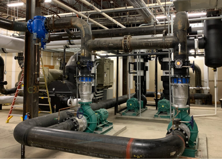 Industrial mechanical room featuring two green centrifugal water pumps, large black piping loops, blue control valves, and background HVAC chiller equipment.