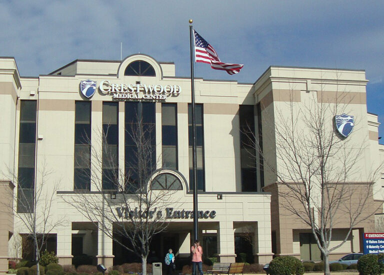 Exterior facade of Crestwood Medical Center featuring the corporate logo, a covered Visitor's Entrance, and an American flag flying on a central flagpole.