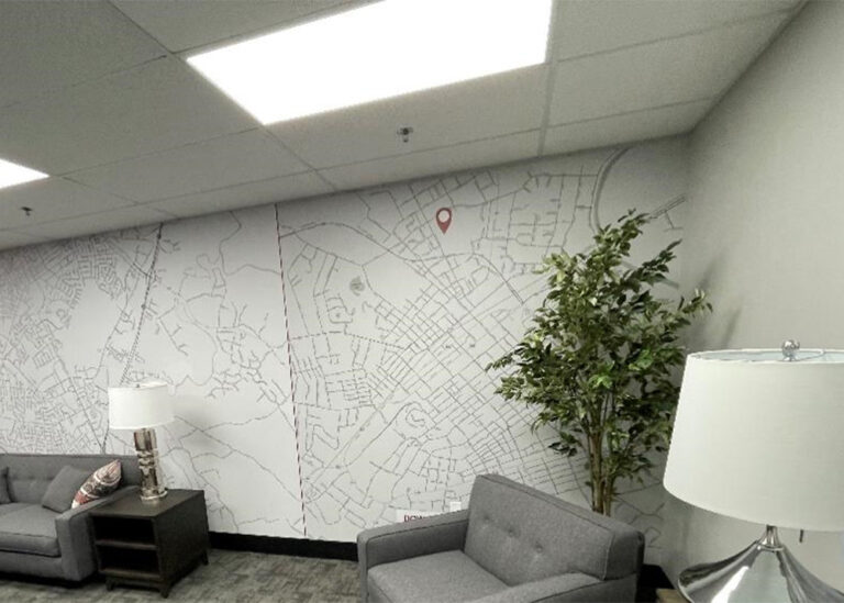 A modern office lounge area featuring grey upholstered seating, table lamps, a potted plant, and a full-wall street map mural marked with a red location pin.