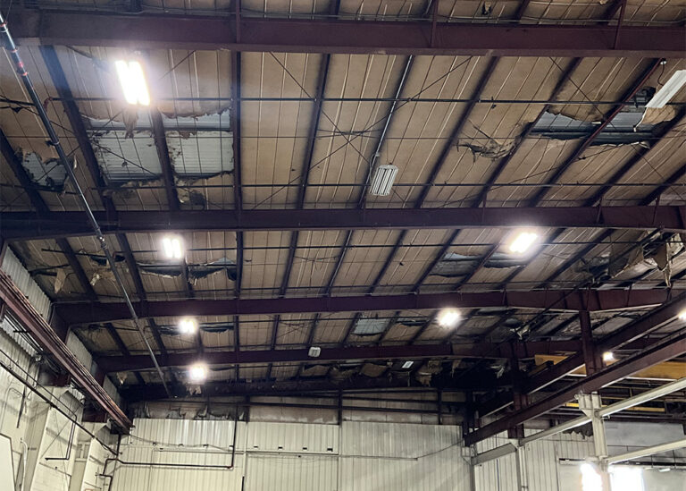 The interior ceiling of an industrial warehouse showing significantly damaged, peeling roof insulation and old high-bay lighting fixtures.