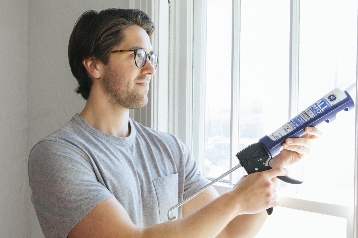 A homeowner uses a caulking gun to apply sealant along a residential window frame to reduce drafts and improve home air sealing.