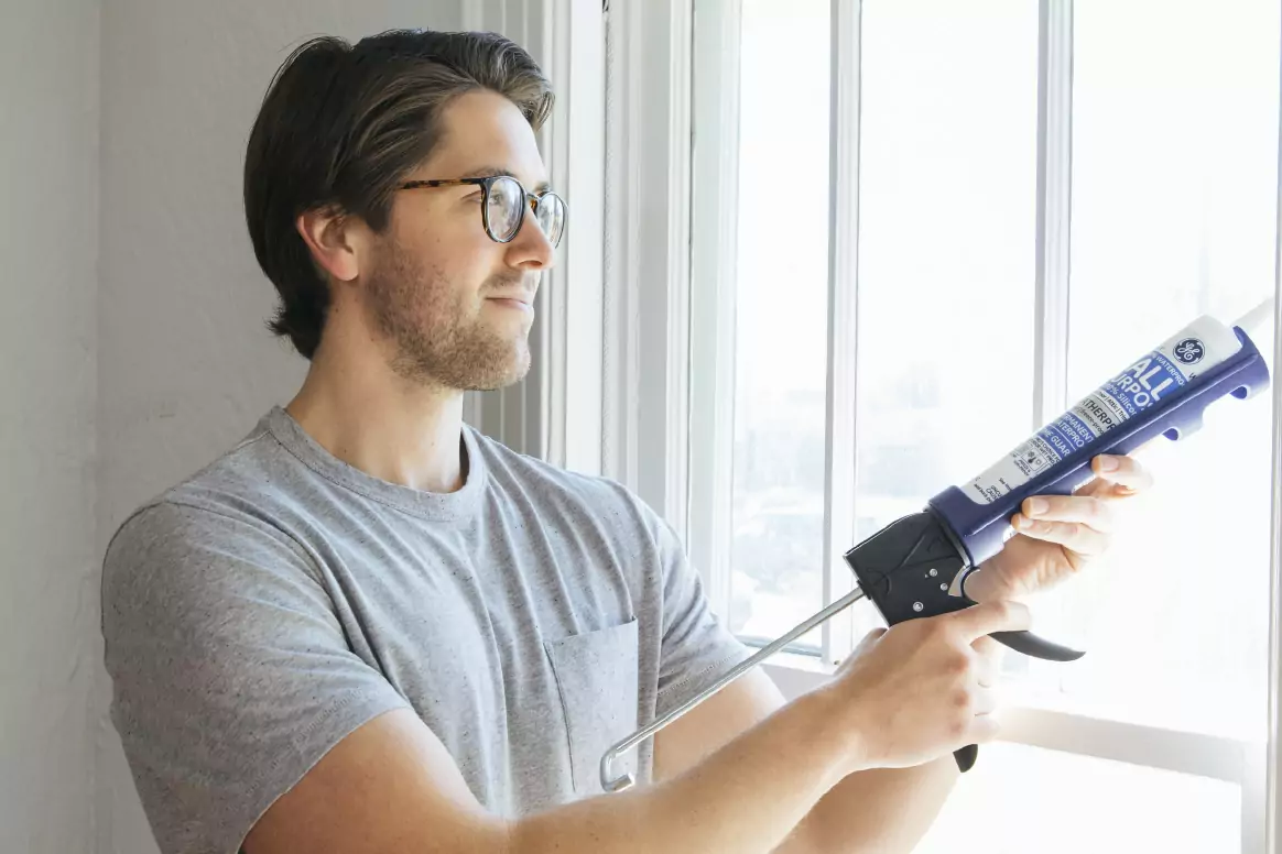 A homeowner uses a caulking gun to apply sealant along a residential window frame to reduce drafts and improve home air sealing.