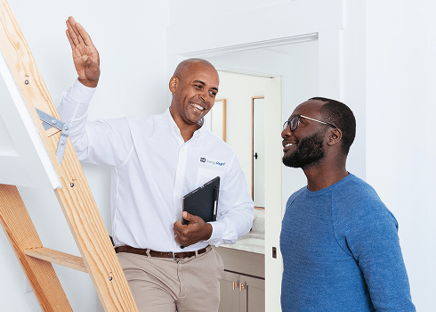 A smiling TVA Energy Expert holding a digital tablet and gesturing toward a pull-down wooden ladder while conducting a home energy assessment for a resident.
