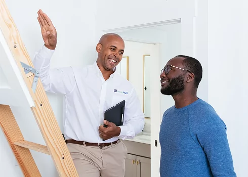 A smiling TVA Energy Expert holding a digital tablet and gesturing toward a pull-down wooden ladder while conducting a home energy assessment for a resident.