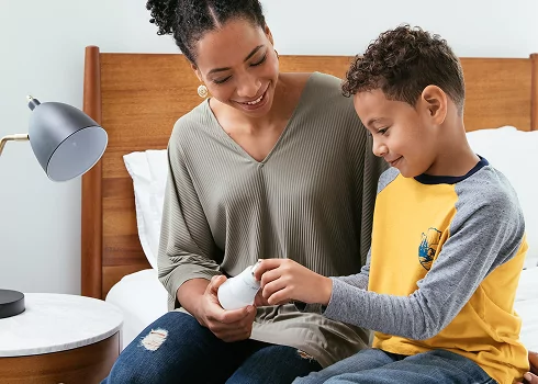 A mother and her young son sitting on a bed together examining an energy-efficient LED lightbulb next to a bedside lamp.