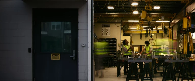 An open doorway reveals a brightly lit manufacturing facility where workers in high-visibility vests operate equipment under energy-efficient industrial overhead lighting.