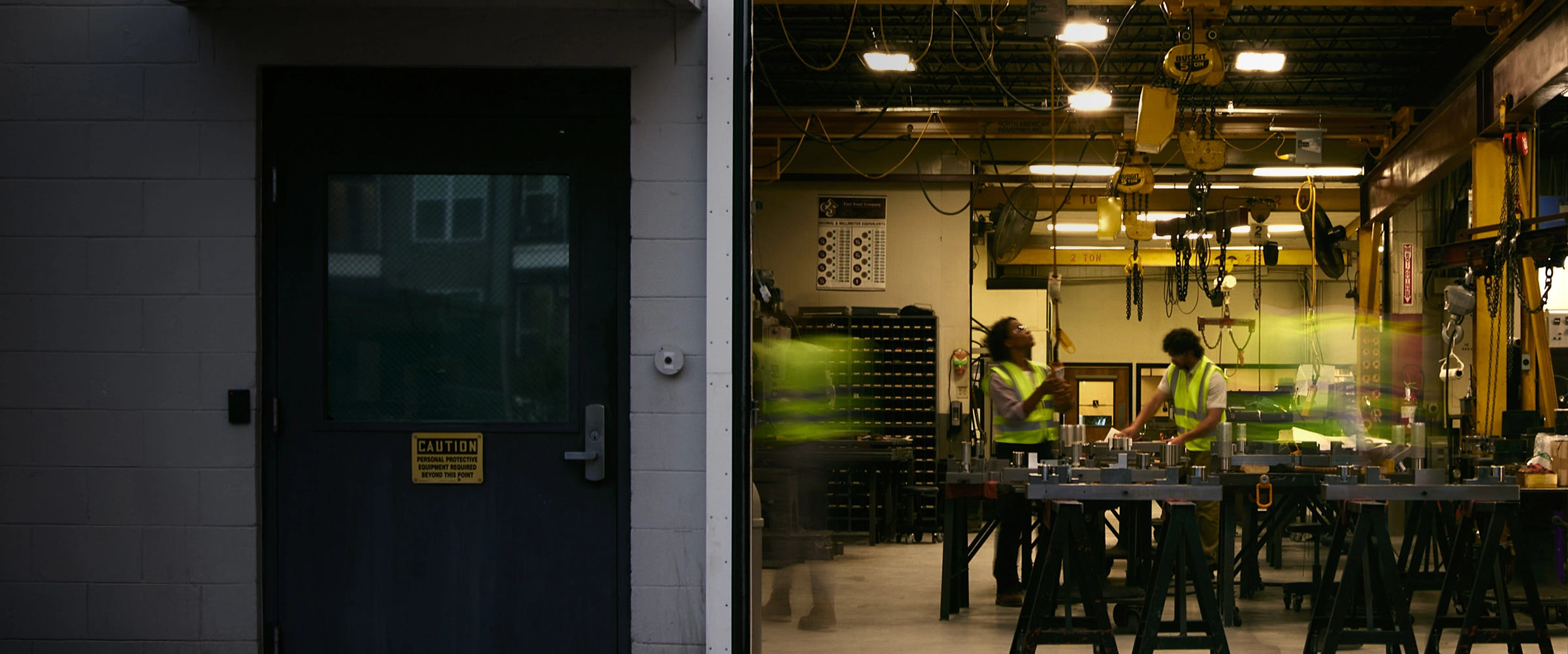 An open doorway reveals a brightly lit manufacturing facility where workers in high-visibility vests operate equipment under energy-efficient industrial overhead lighting.