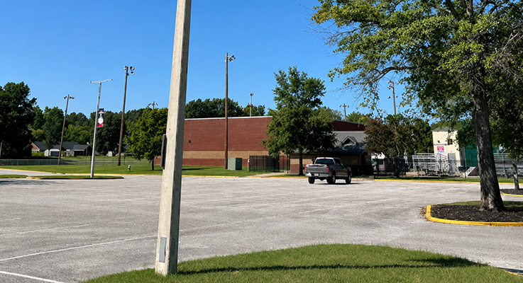 Community center parking lot with a brick building, mature trees, and stadium light poles, ideal for future energy-efficient upgrades.