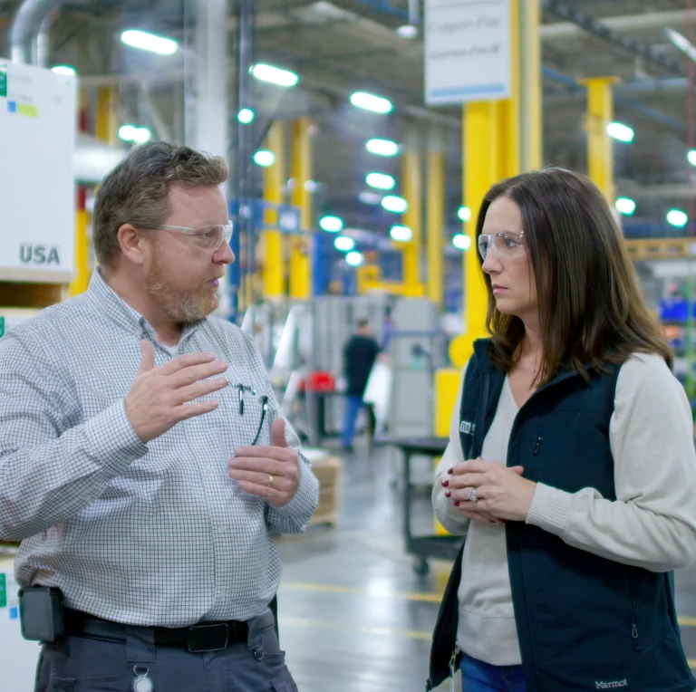 A TVA Energy Expert wearing protective eyewear gestures while discussing industrial energy-efficient upgrades with a facility representative on a factory floor.